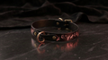 A black leather collar with a gold-colored buckle and a floral design on the strap is displayed on a dark surface.