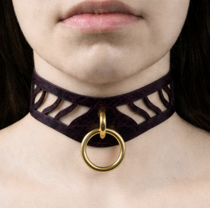 A close-up of a person's neck and chest area, featuring a dark purple choker necklace with a gold ring and a cutout design.