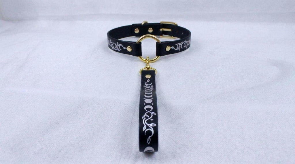 A black collar with a gold-colored buckle and a decorative handle attached to it, resting on a white surface.