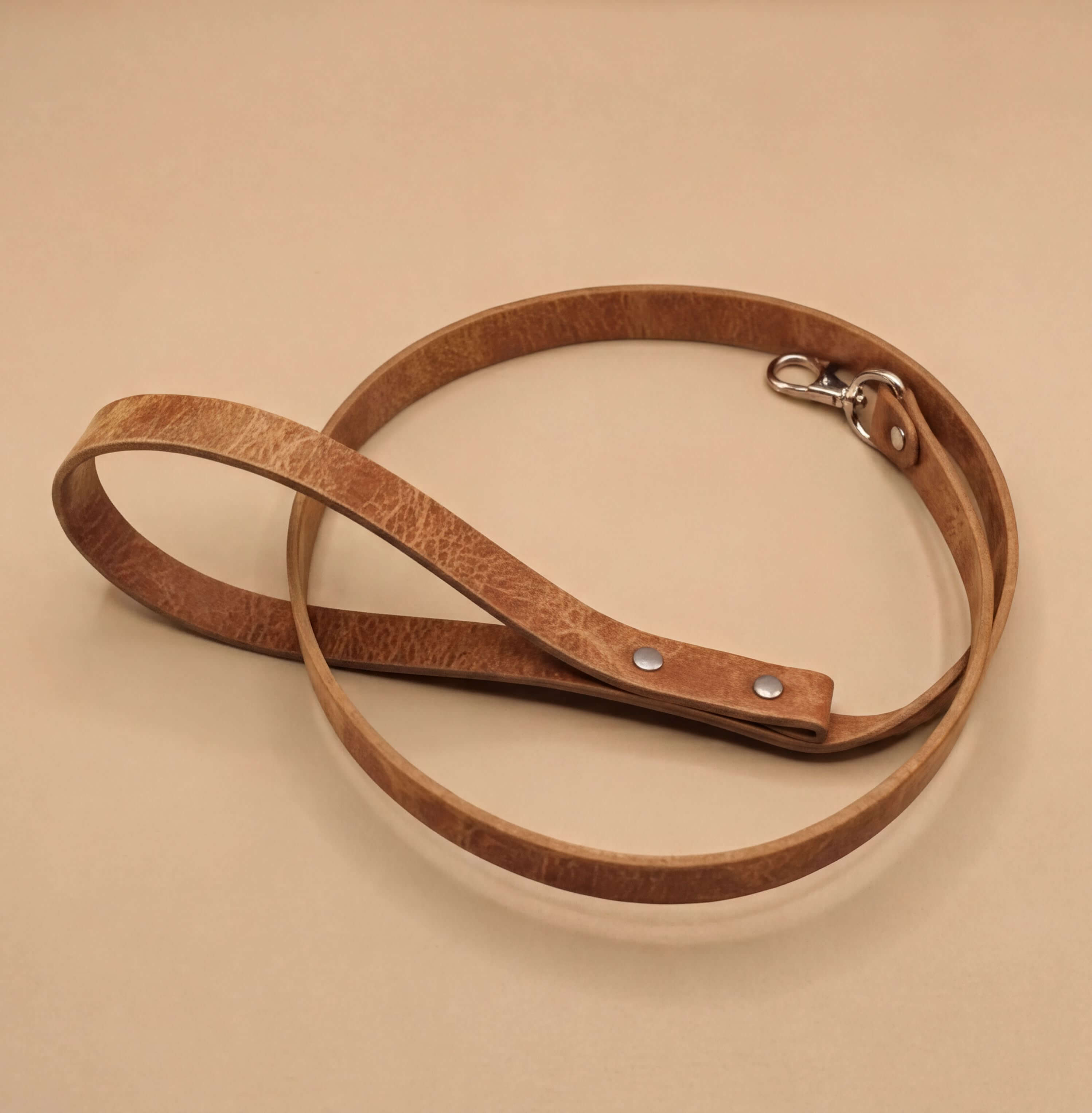 A brown leather dog leash with a metal clasp and buckle is shown on a beige surface.