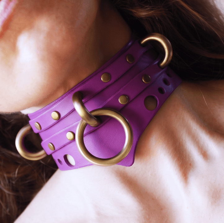 A close-up of a person's neck wearing a purple leather choker necklace with gold-colored metal rings.