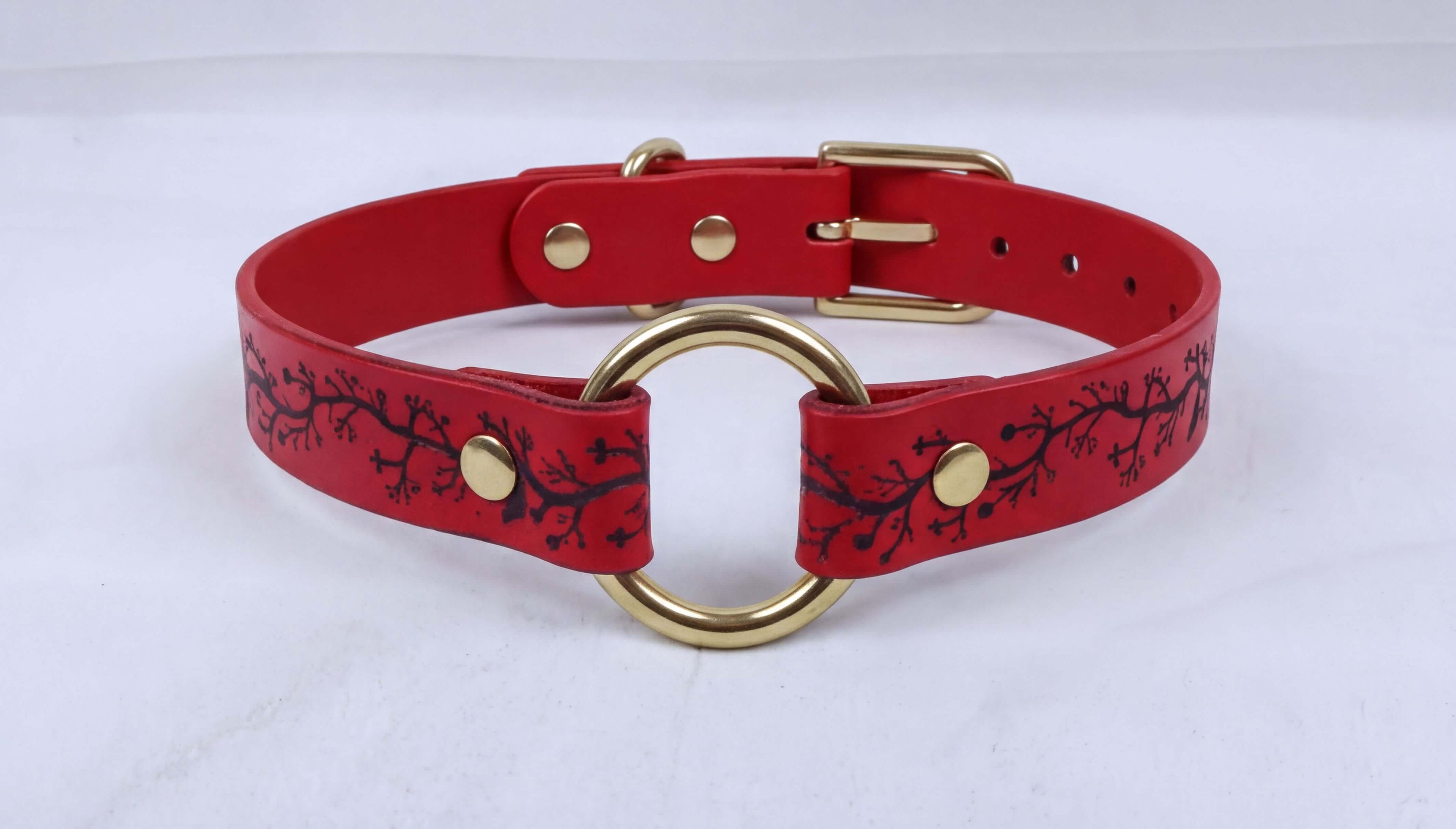 A red leather choker collar with a gold ring and a black branch design.