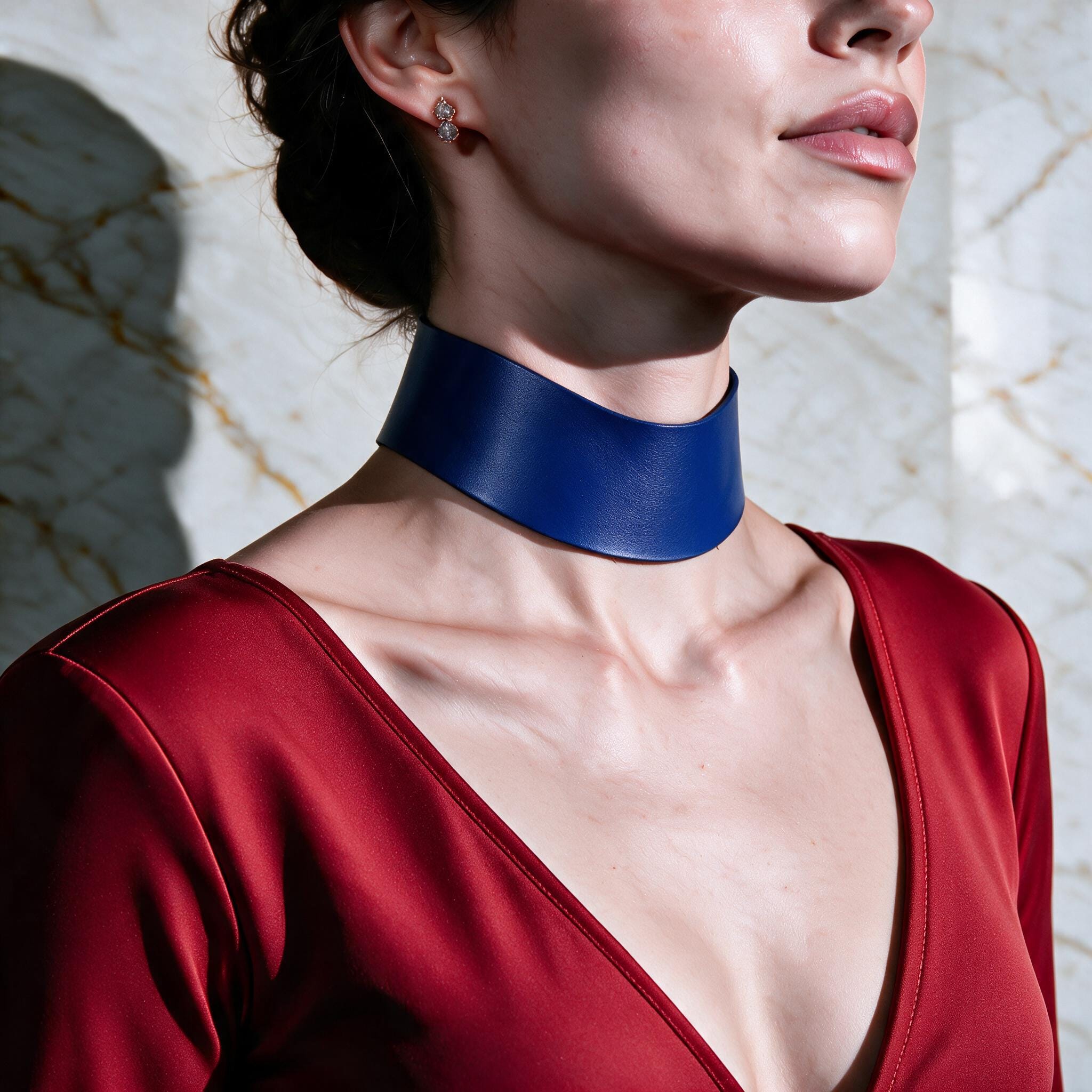 A woman wearing a red dress and a blue choker necklace stands against a marble wall.