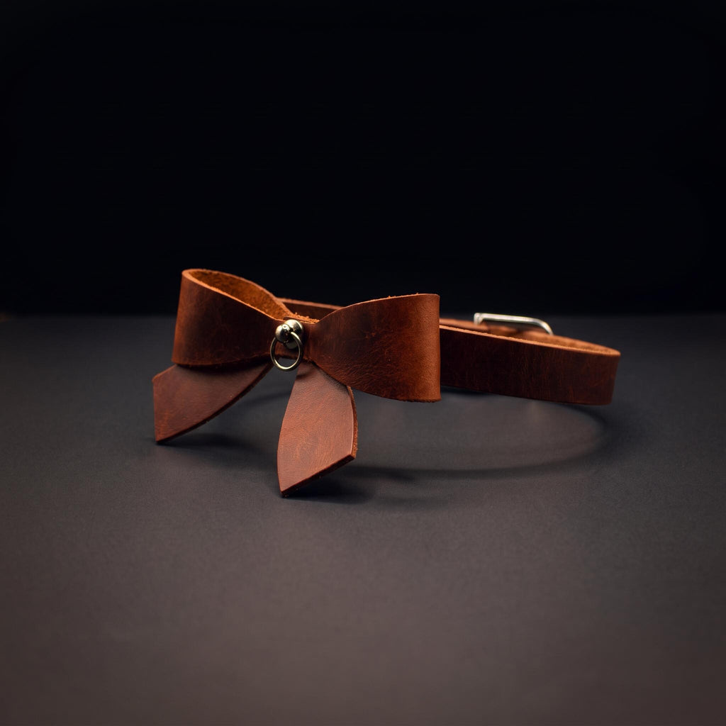 A brown leather bow tie with a metal clasp is displayed against a dark background.