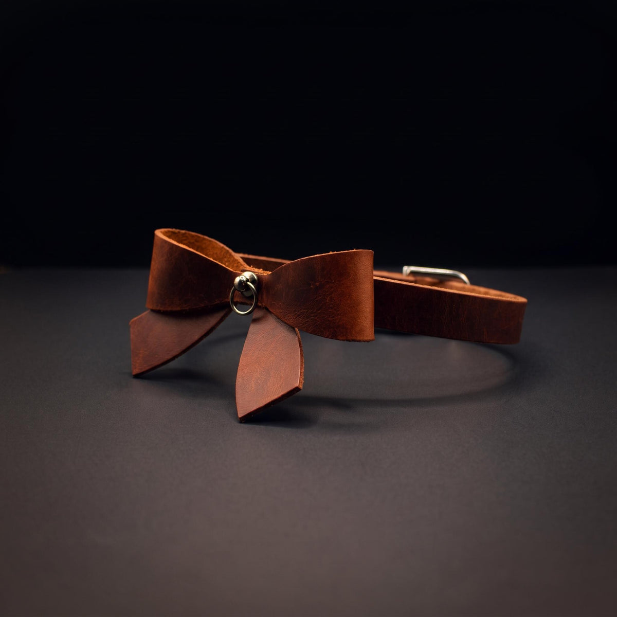 A brown leather bow tie with a metal clasp is displayed against a dark background.