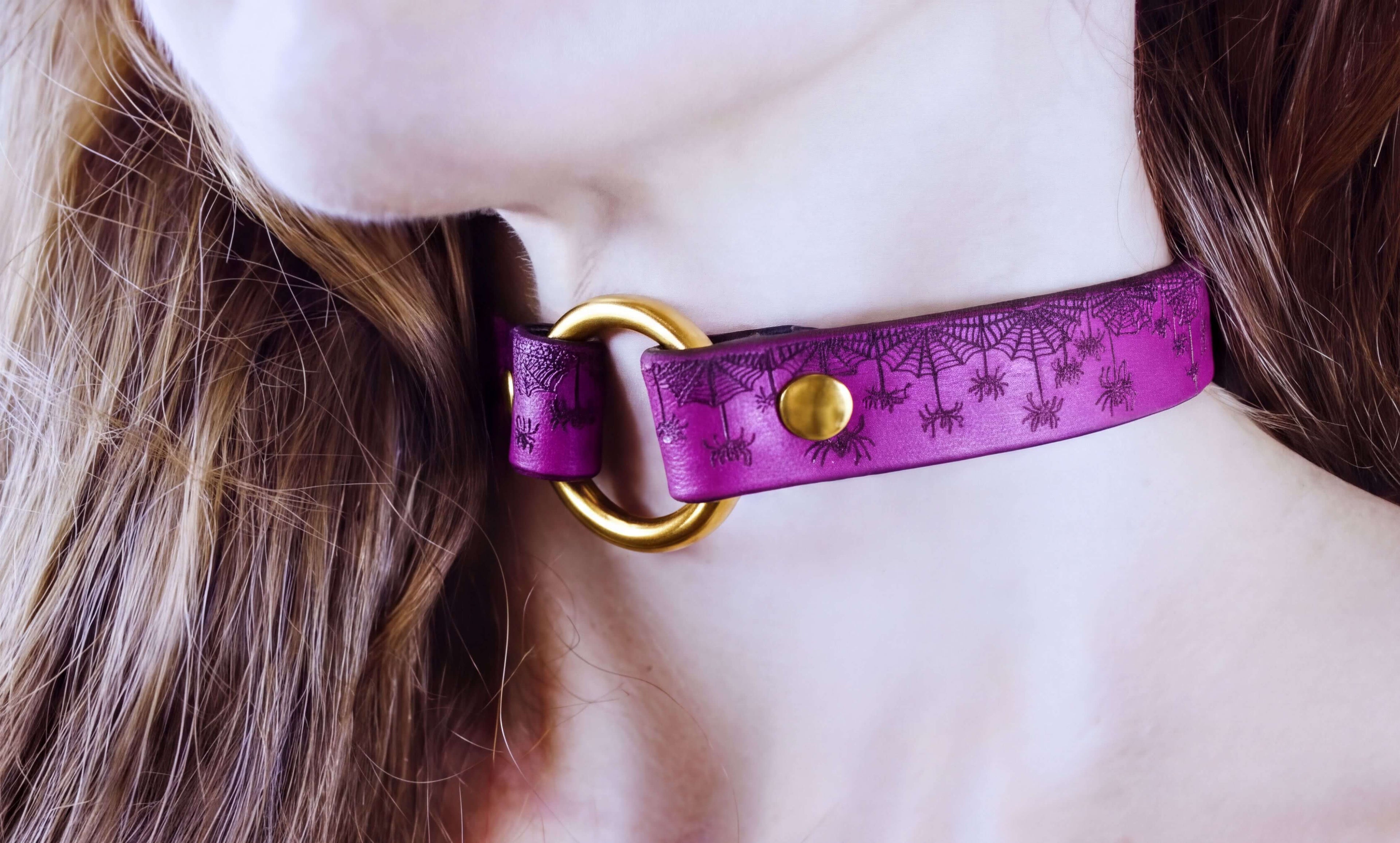A close-up of a person's neck and chin wearing a purple choker necklace with a gold-colored metal ring.