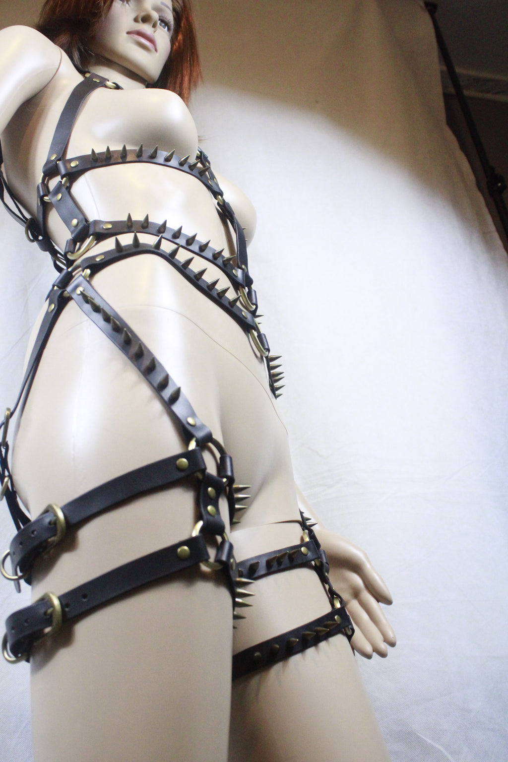 A mannequin wearing a leather strap ensemble with spikes, posing on a white surface.