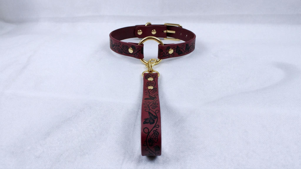 A burgundy leather collar with a gold buckle and a black floral pattern on the strap is displayed on a white surface.