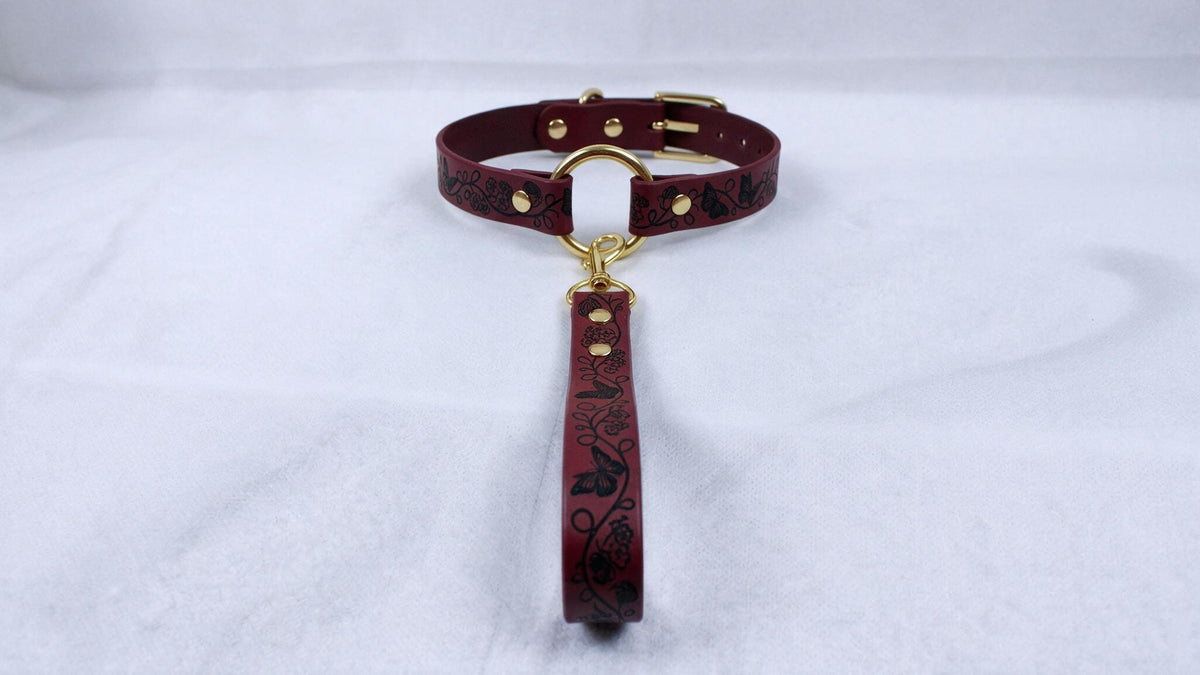 A burgundy leather collar with a gold buckle and a black floral pattern on the strap is displayed on a white surface.