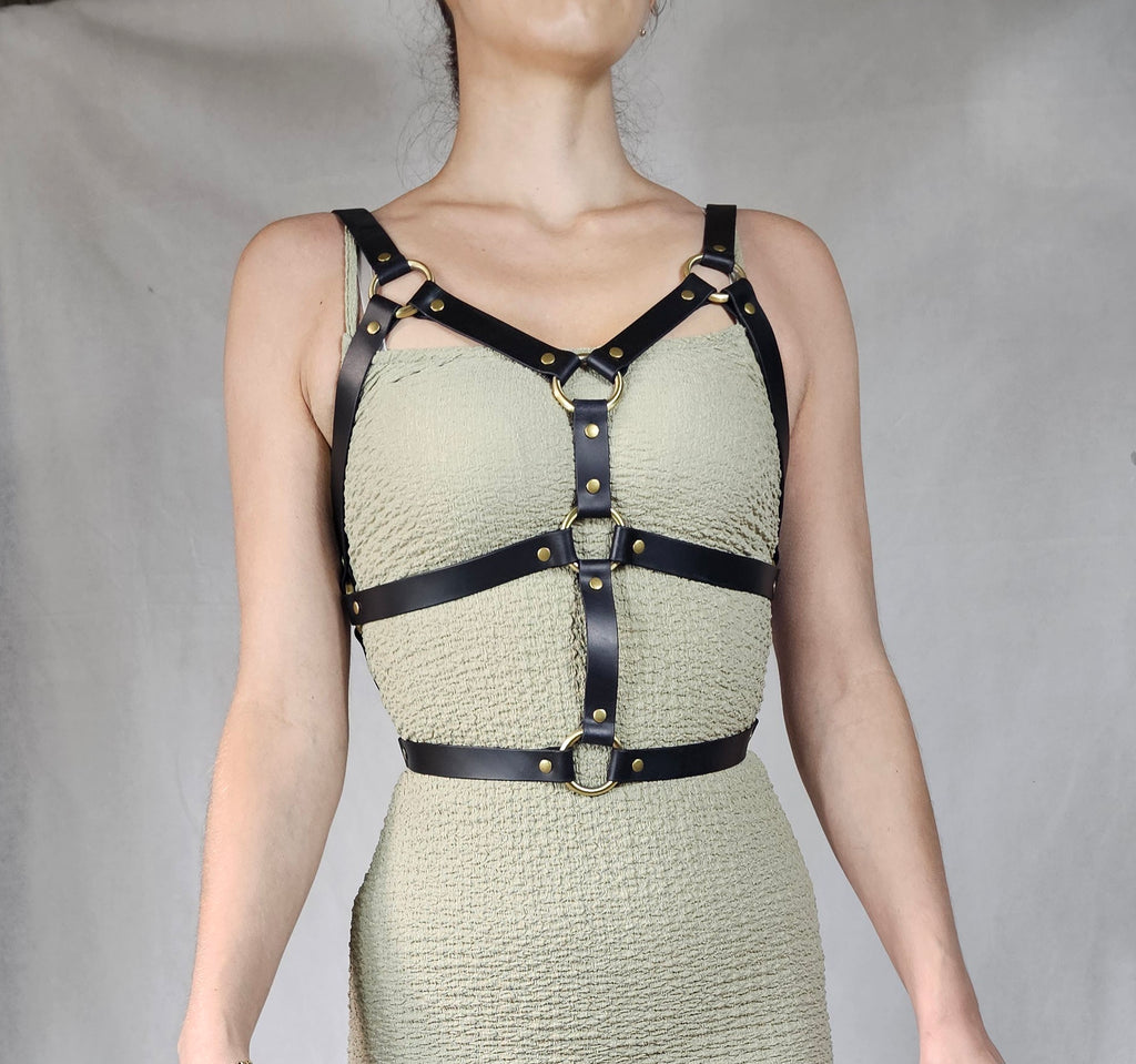 A woman wearing a beige dress with a leather harness and straps, posing against a plain background.
