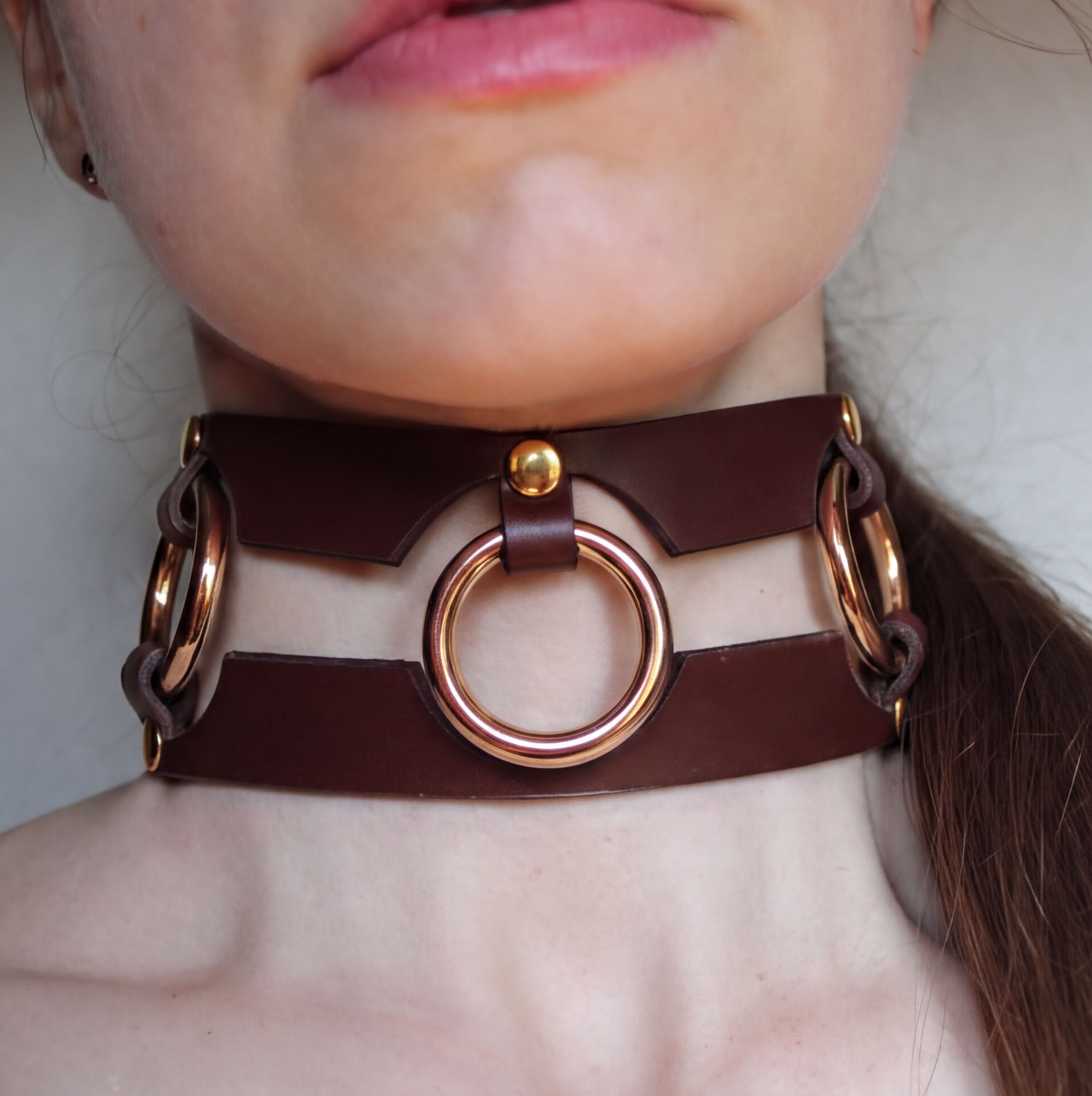 A close-up of a person's neck and chin wearing a brown leather choker necklace with three circular rings.