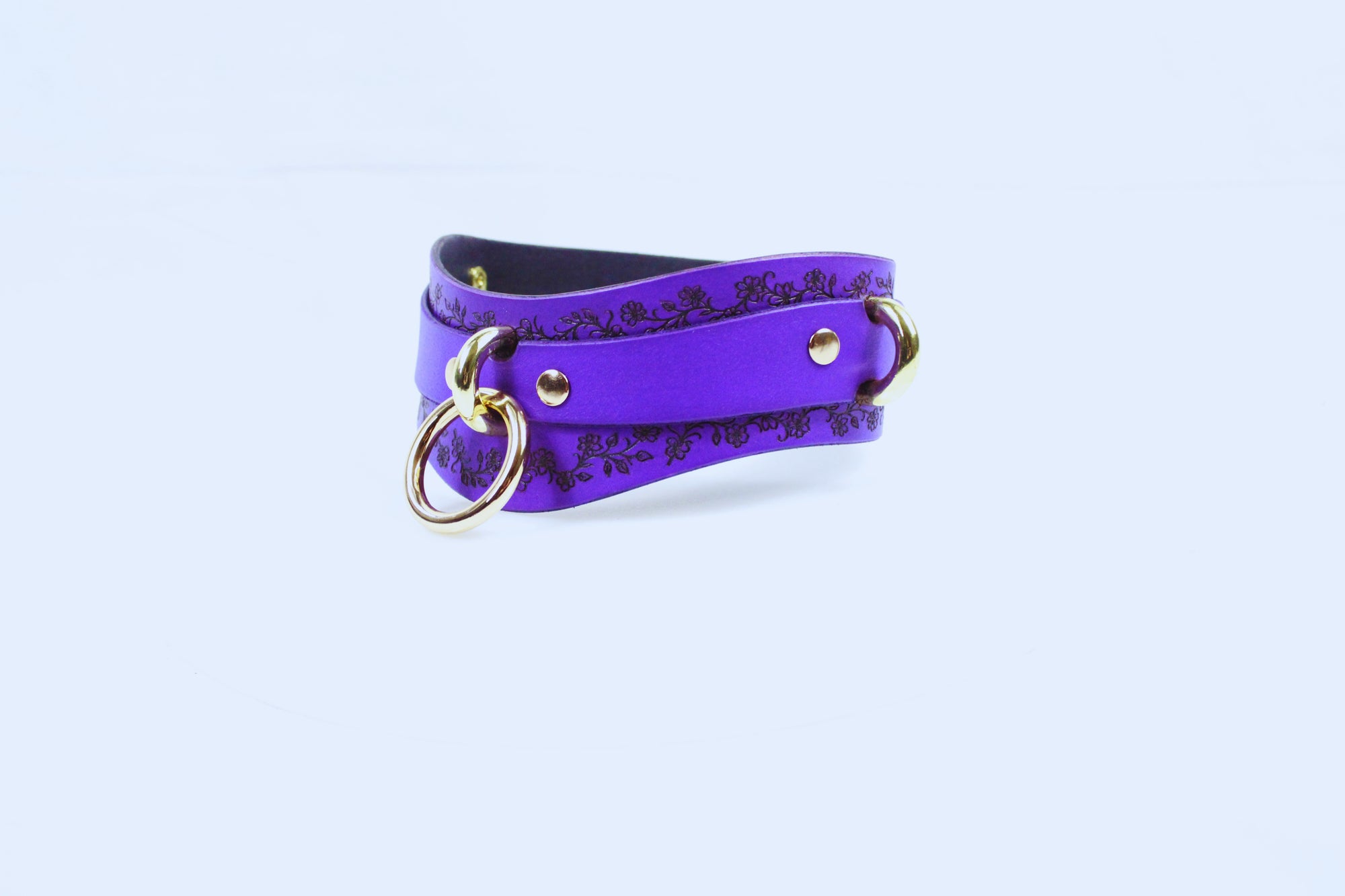 A purple leather choker with a floral pattern and gold-colored metal rings is shown against a white background.