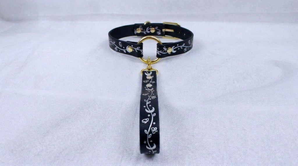 A black collar with a gold-colored metal clasp and a decorative metal tag hanging from it, placed on a white surface.