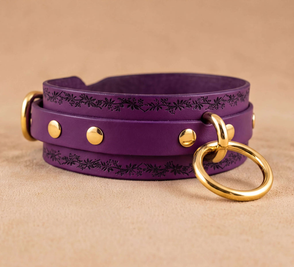 A purple leather choker necklace with gold-colored metal rings and studs, resting on a beige surface.
