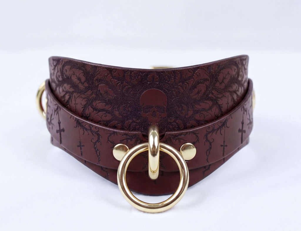 A brown leather choker necklace with a gold-colored metal ring and a skull design on the front.