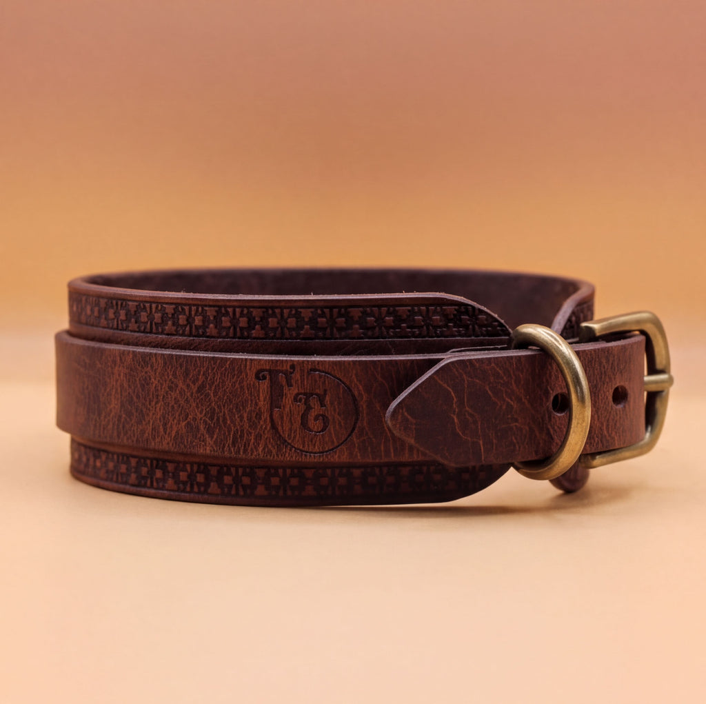 A brown leather belt with a metal buckle, featuring a decorative pattern around the buckle.