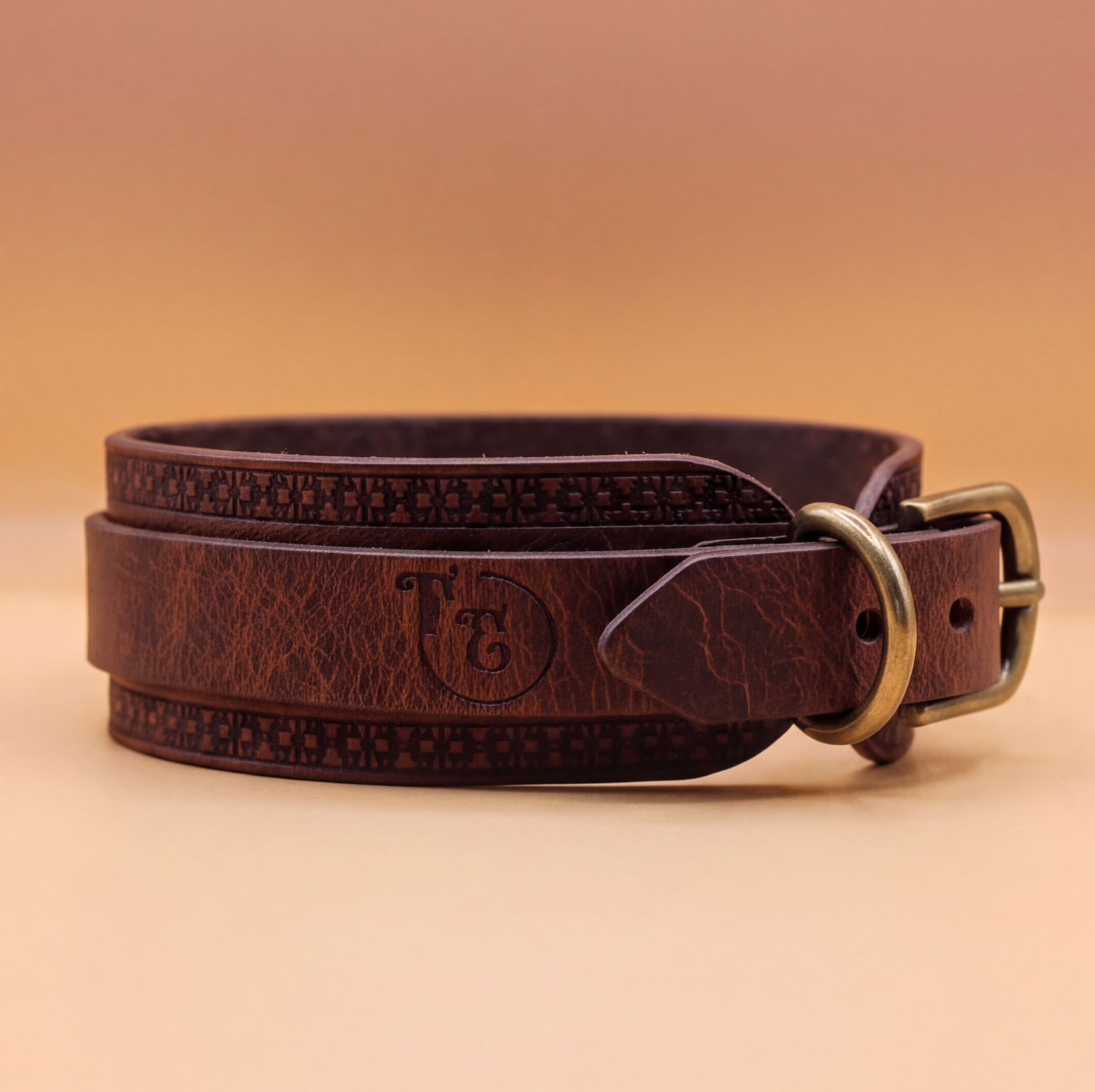A brown leather belt with a metal buckle, featuring a decorative pattern around the buckle.