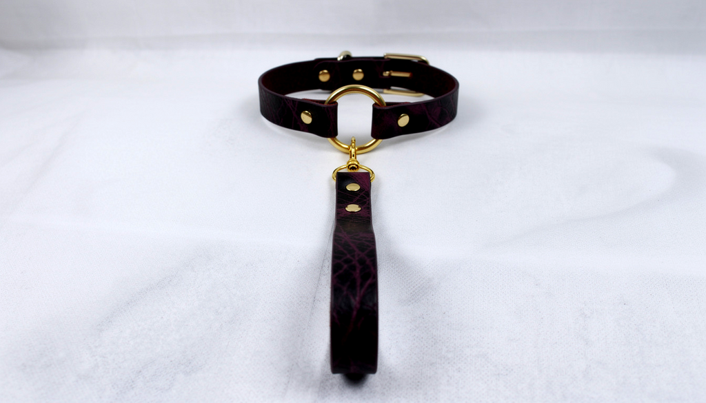 A dark purple leather collar with a gold-colored buckle and a long strap attached to it, resting on a white surface.