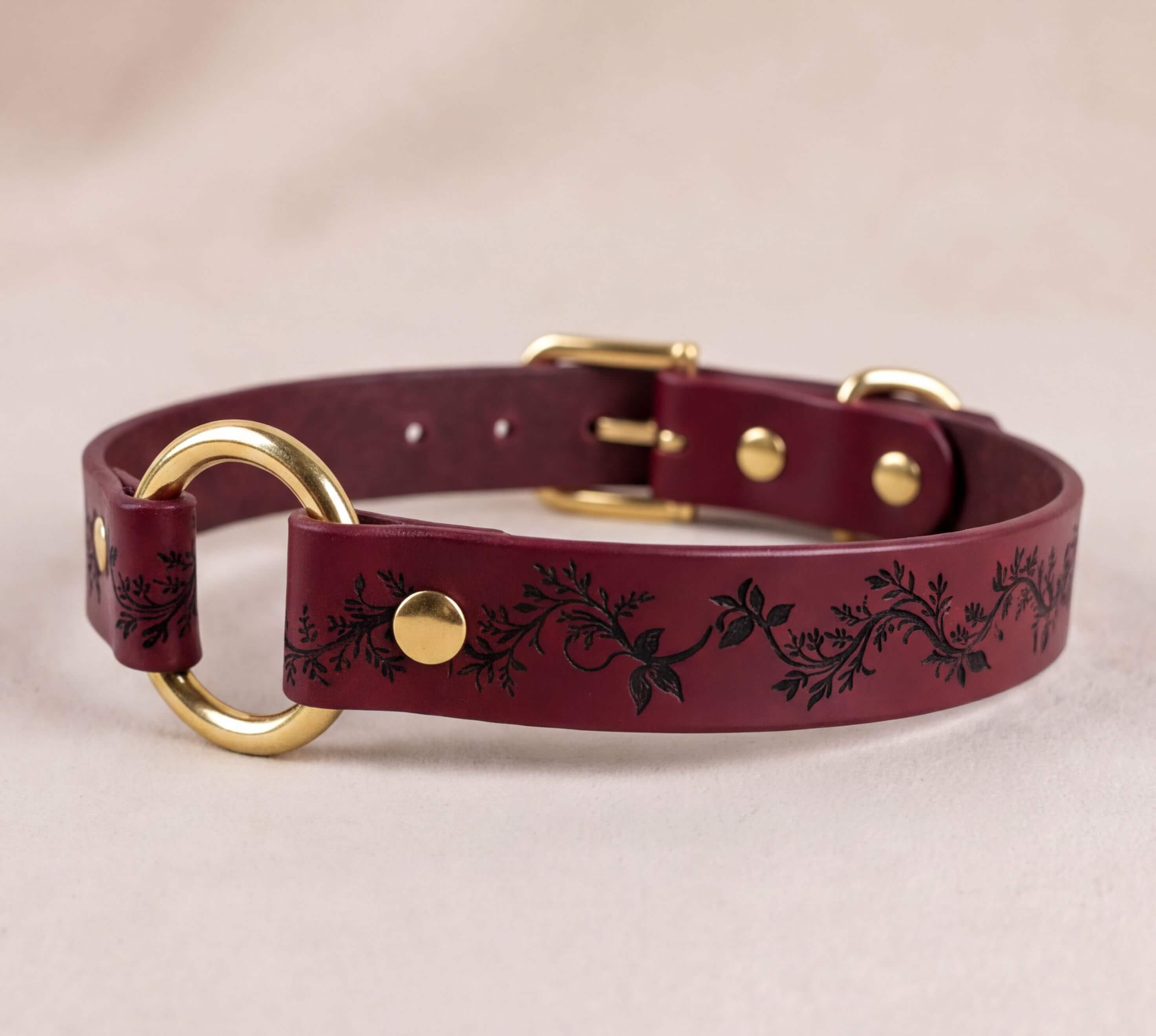 A burgundy leather dog collar with a gold-colored buckle and a decorative leaf pattern.