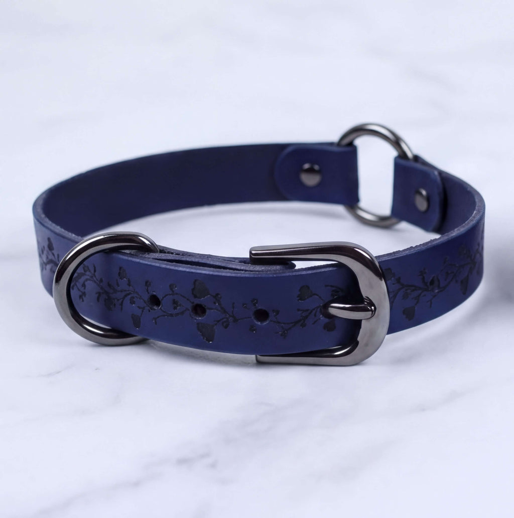 A navy blue leather collar with a metal buckle and two metal rings, resting on a white marble surface.