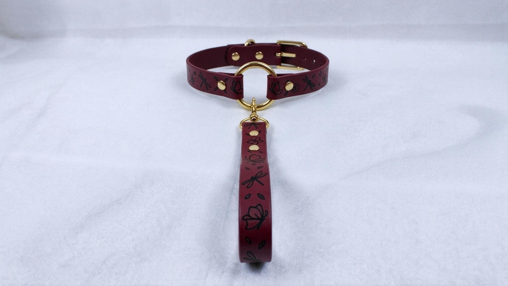 A burgundy leather collar with a gold-colored buckle and a black floral pattern on the strap.