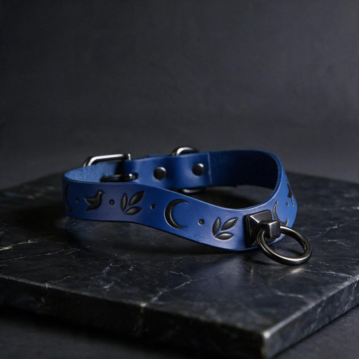 A blue leather collar with a moon and leaf design, featuring a metal ring and buckle, is displayed on a dark surface.
