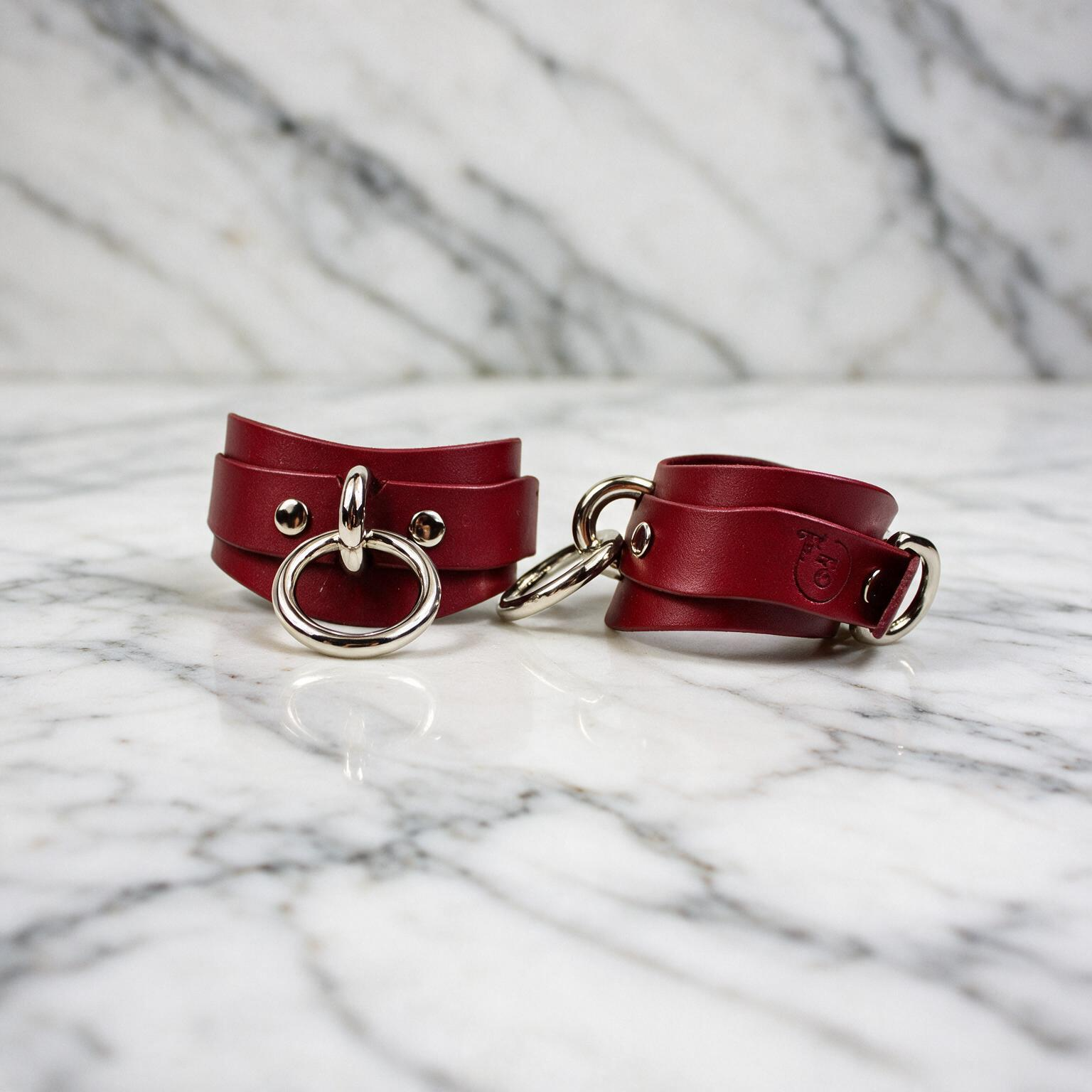 A pair of red leather wrist straps with metal rings and buckles, placed on a marble surface.