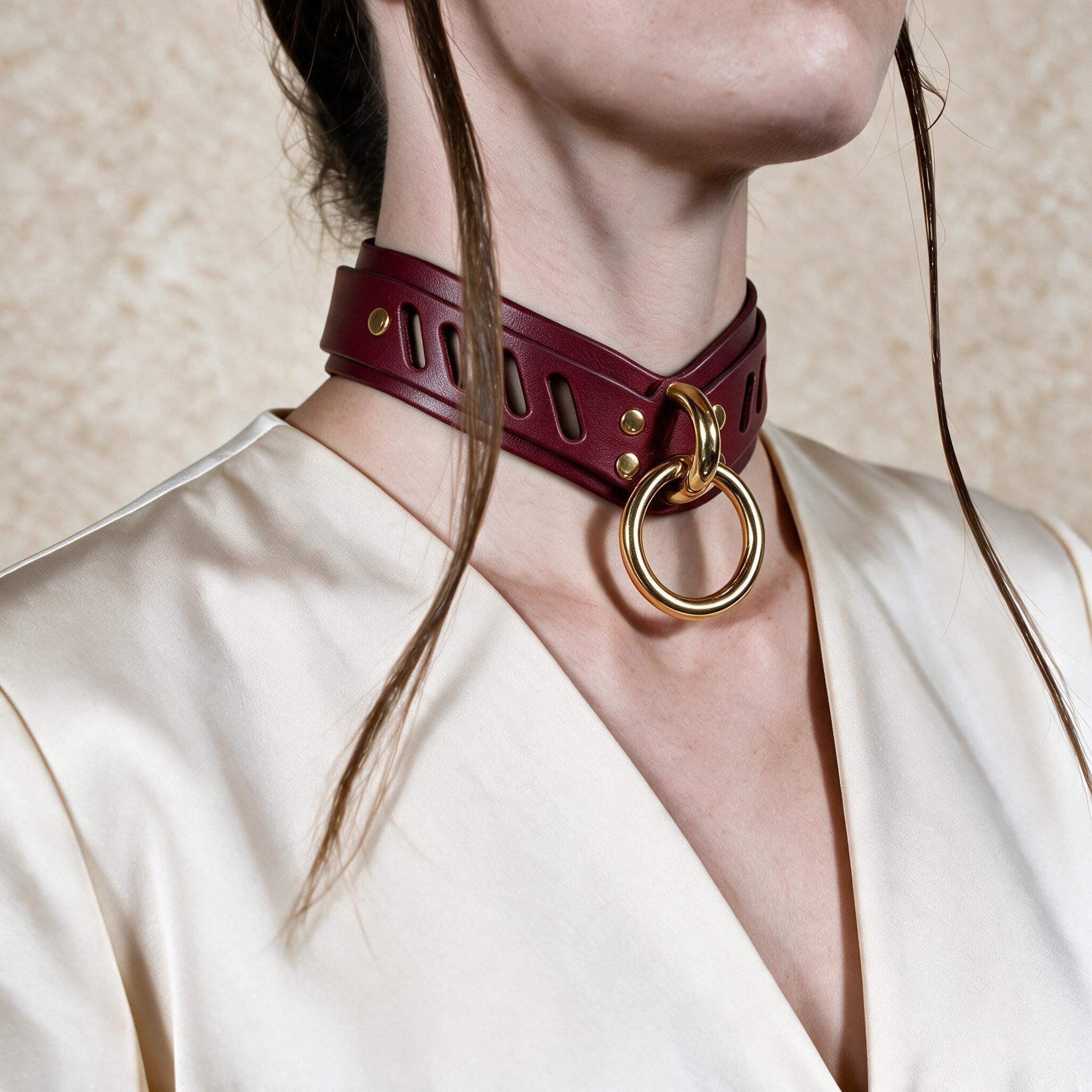 A close-up of a person wearing a burgundy leather choker necklace with a gold ring, against a plain background.