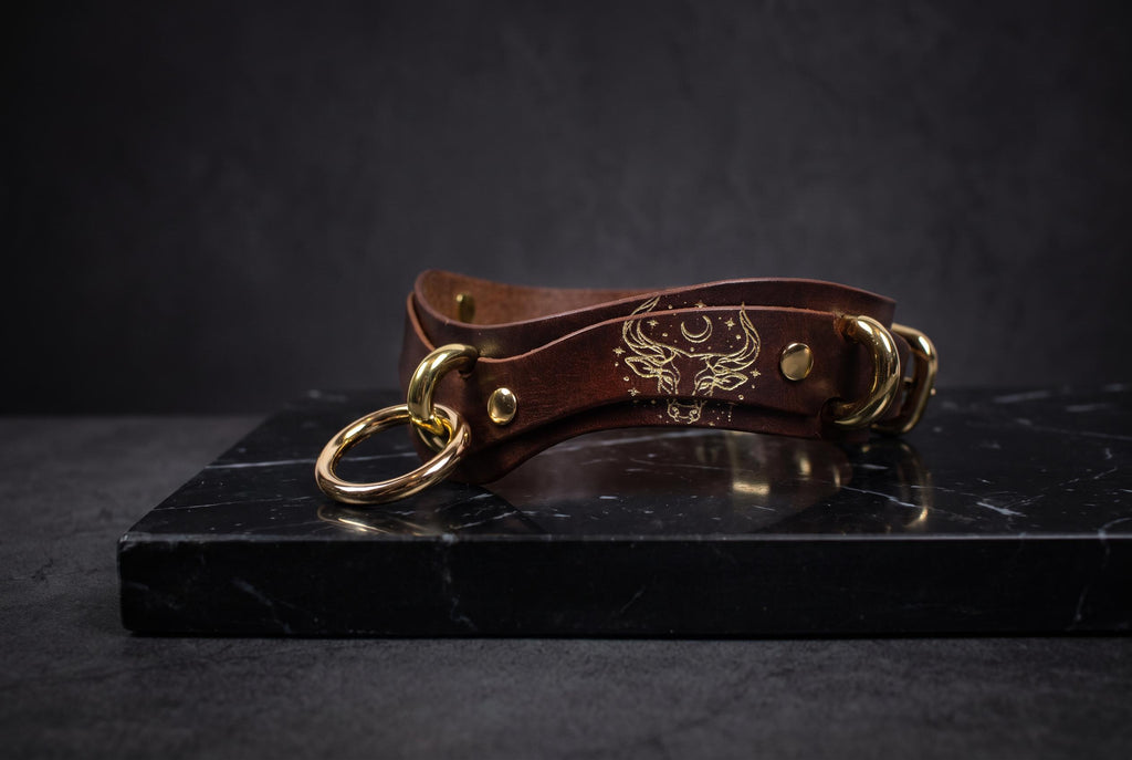 A brown leather choker necklace with gold-colored metal rings and a decorative emblem on the clasp is displayed on a black marble surface.