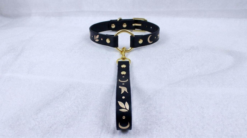 A black leather collar with gold accents and a dangling handle is displayed on a white surface.