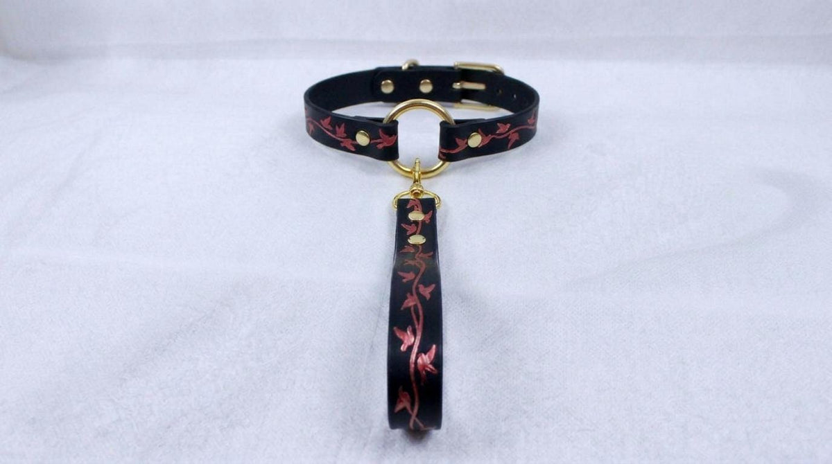 A black leather collar with a gold-colored metal ring and a red floral pattern on the strap.