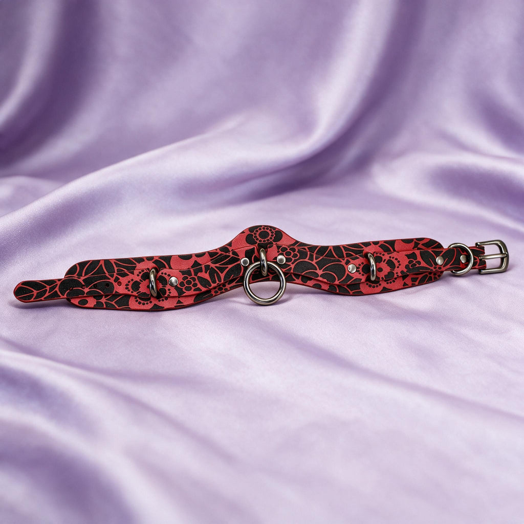 A red and black floral choker necklace with metal rings and a metal buckle is displayed on a purple fabric background.