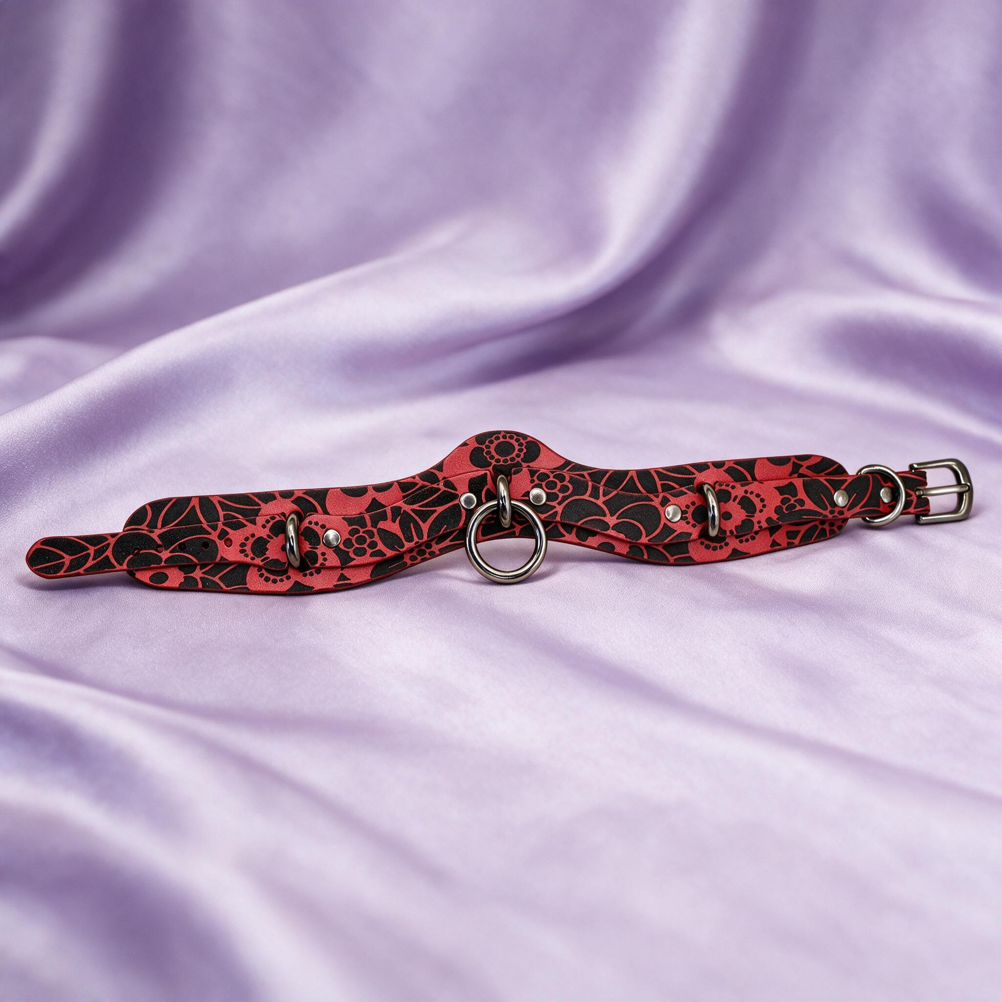 A red and black floral choker necklace with metal rings and a metal buckle is displayed on a purple fabric background.