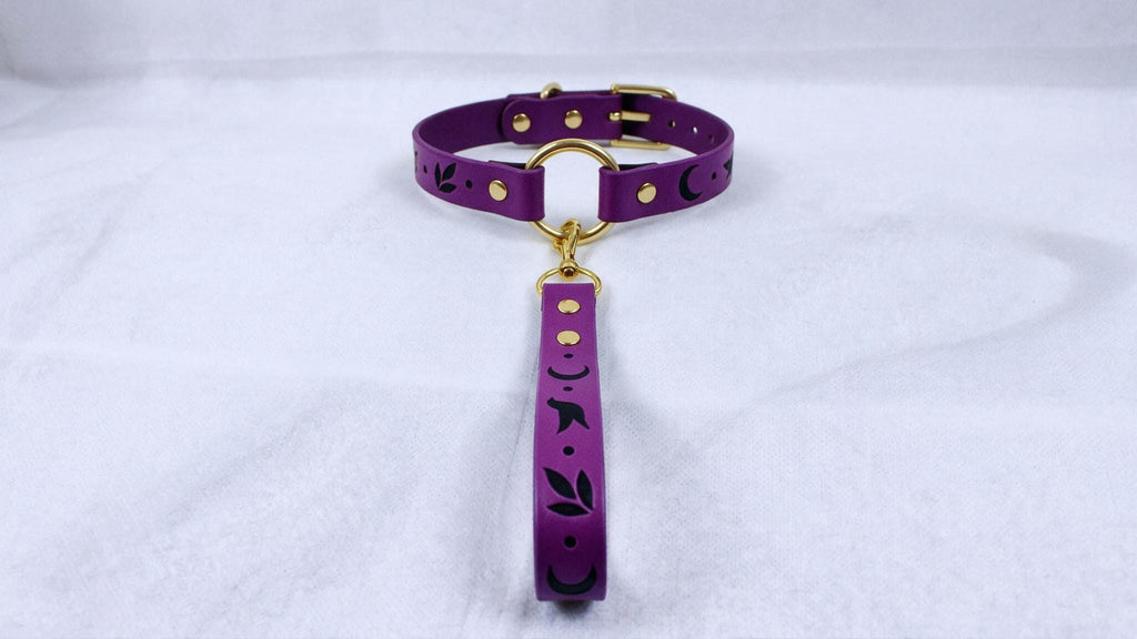 A purple collar with a gold buckle and a black leaf design on it, hanging on a white surface.
