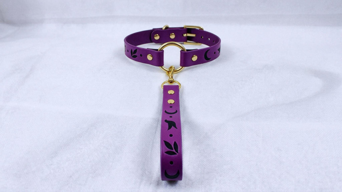 A purple collar with a gold buckle and a black leaf design on it, hanging on a white surface.