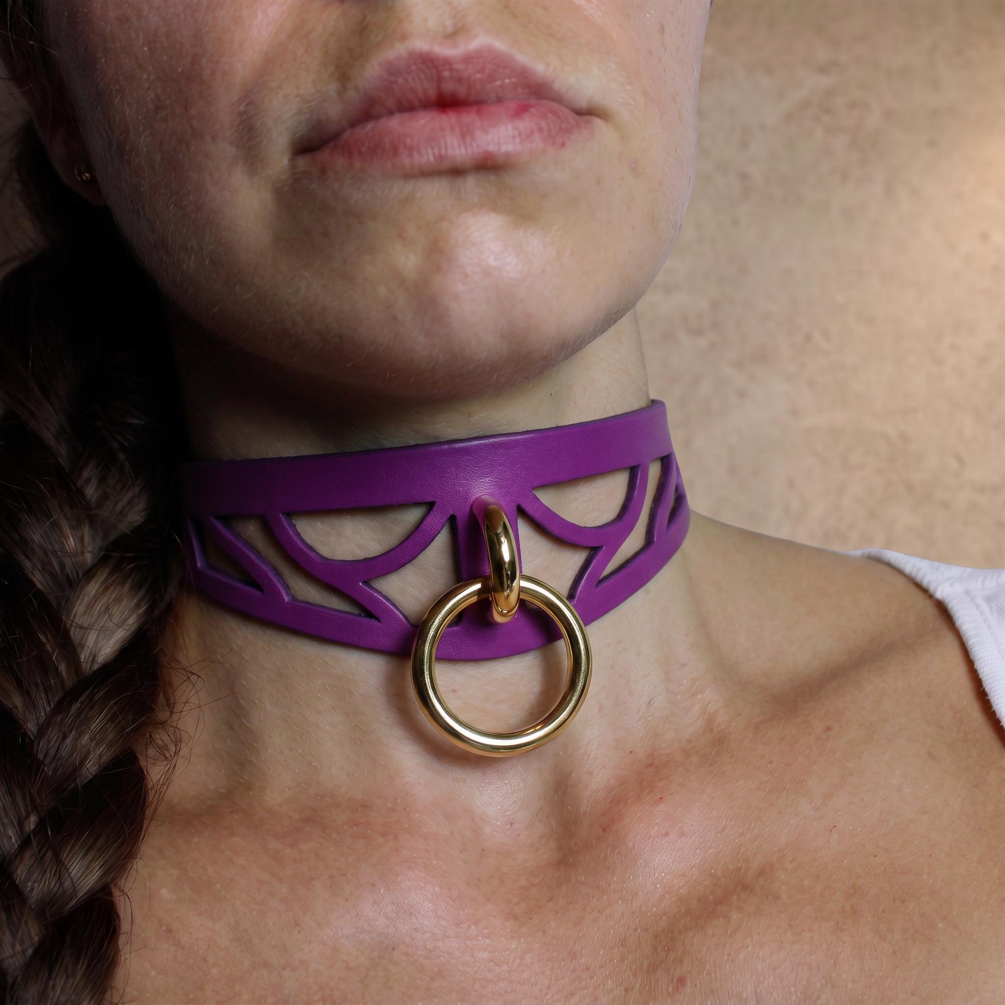 A close-up of a woman's neck and chest area, featuring a purple choker necklace with a gold ring and a gold ring on the collar.