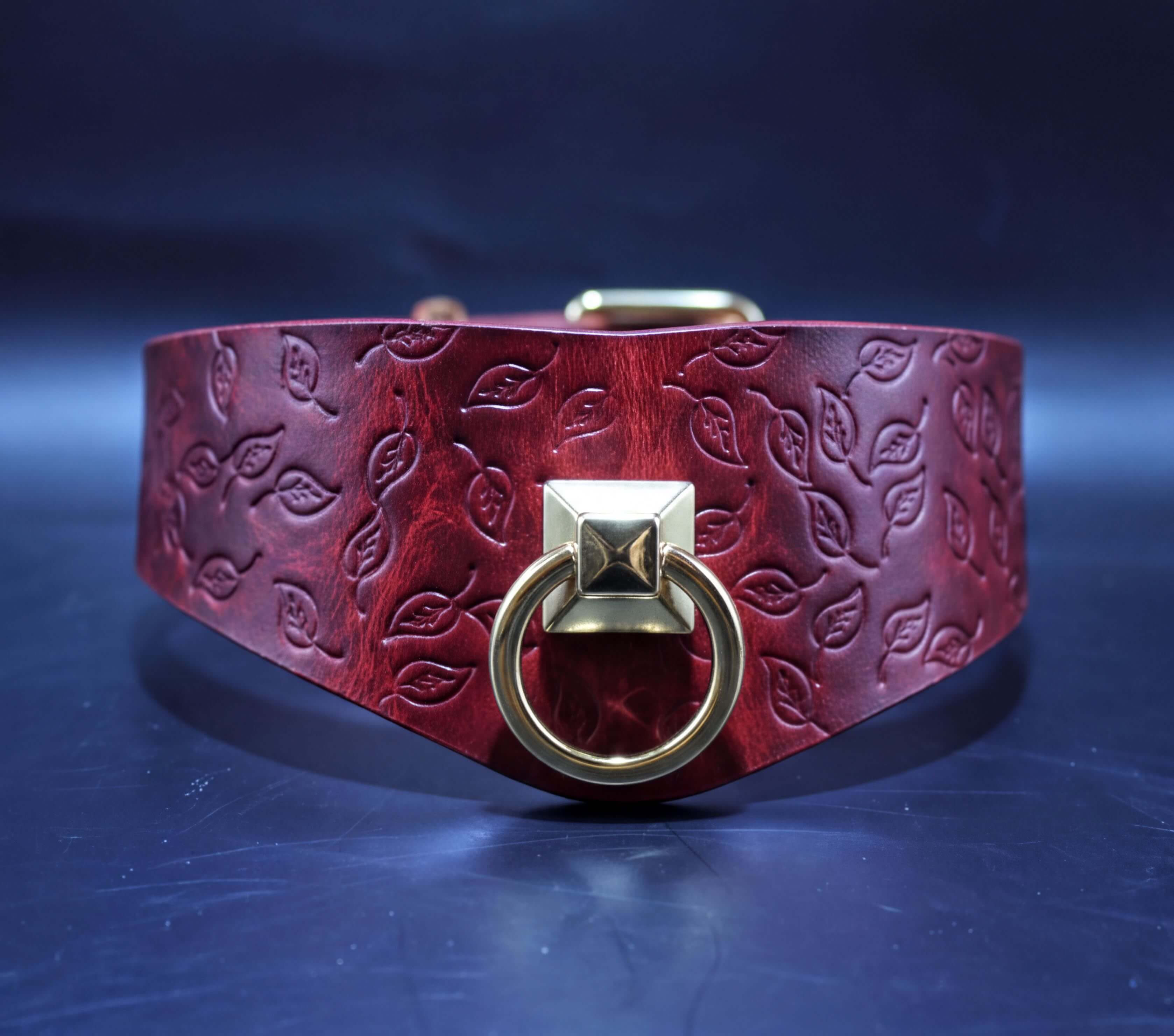A close-up view of a burgundy leather collar with a gold-colored metal ring and a leaf-patterned design.