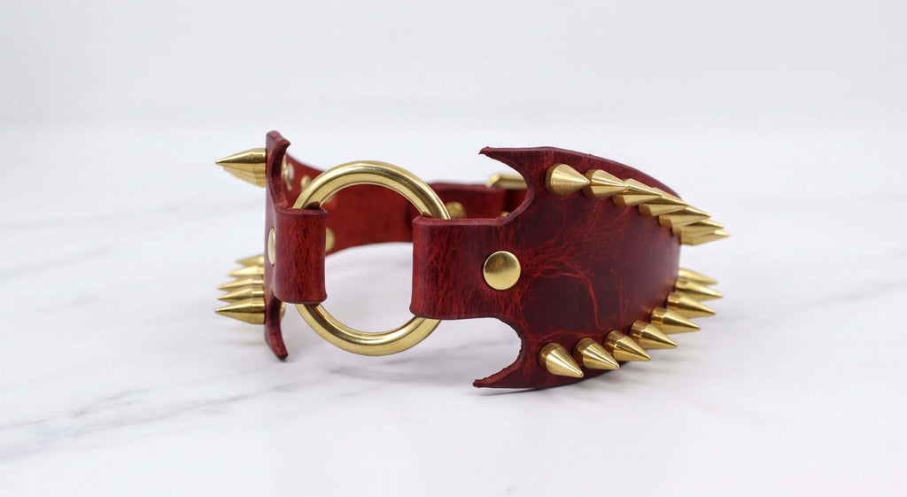 A red leather bracelet with gold spikes protruding from it, set against a white marble background.