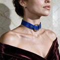A woman wearing a blue choker necklace with a large circular buckle, posing against a marble wall.