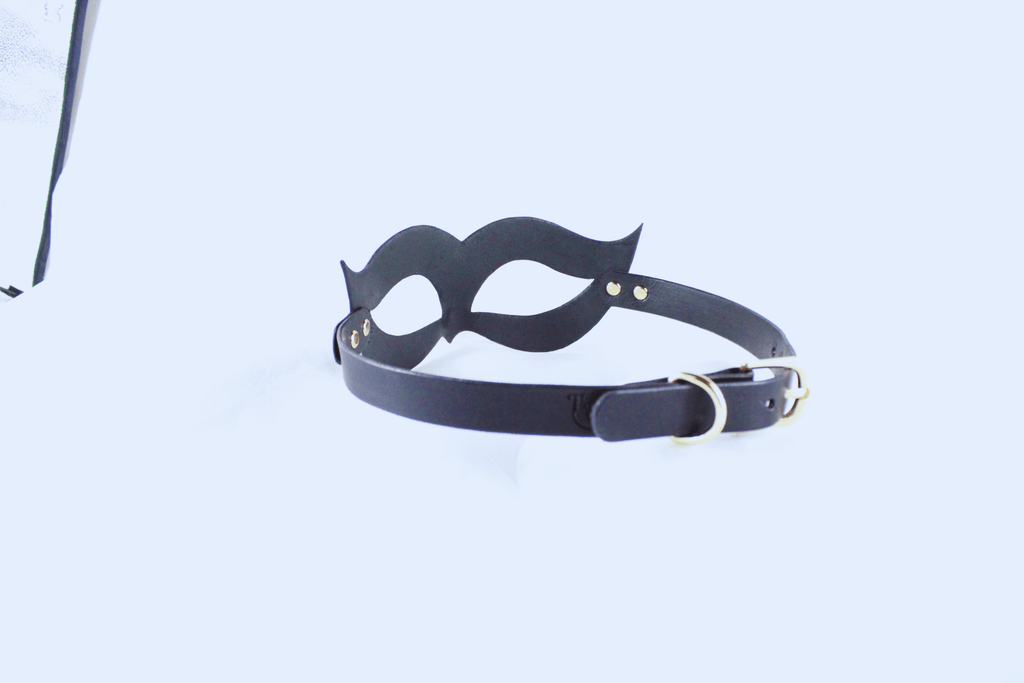 A black mask with a black strap and a gold buckle is shown against a white background.