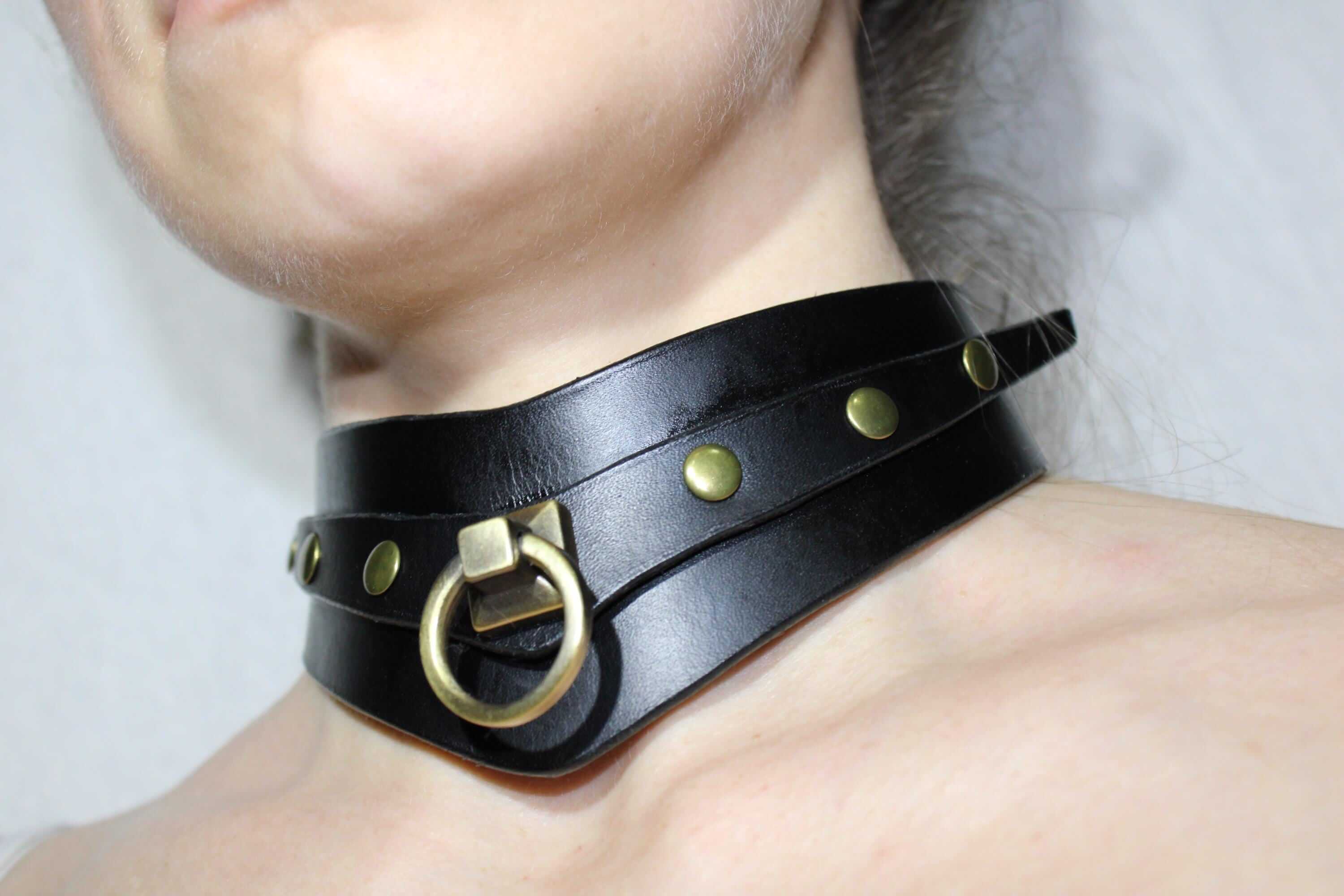 A close-up view of a person's neck wearing a black leather choker necklace with gold-colored studs and a metal ring.