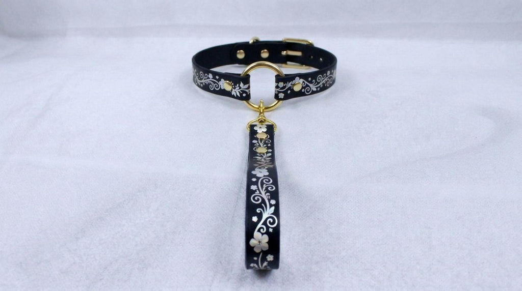 A black collar with a gold-colored buckle and a floral pattern on the strap is displayed against a white background.