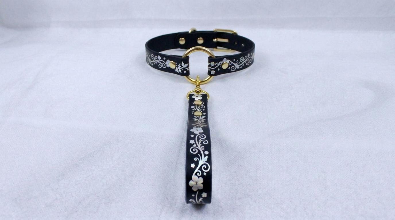 A black collar with a gold-colored buckle and a floral pattern on the strap is displayed against a white background.