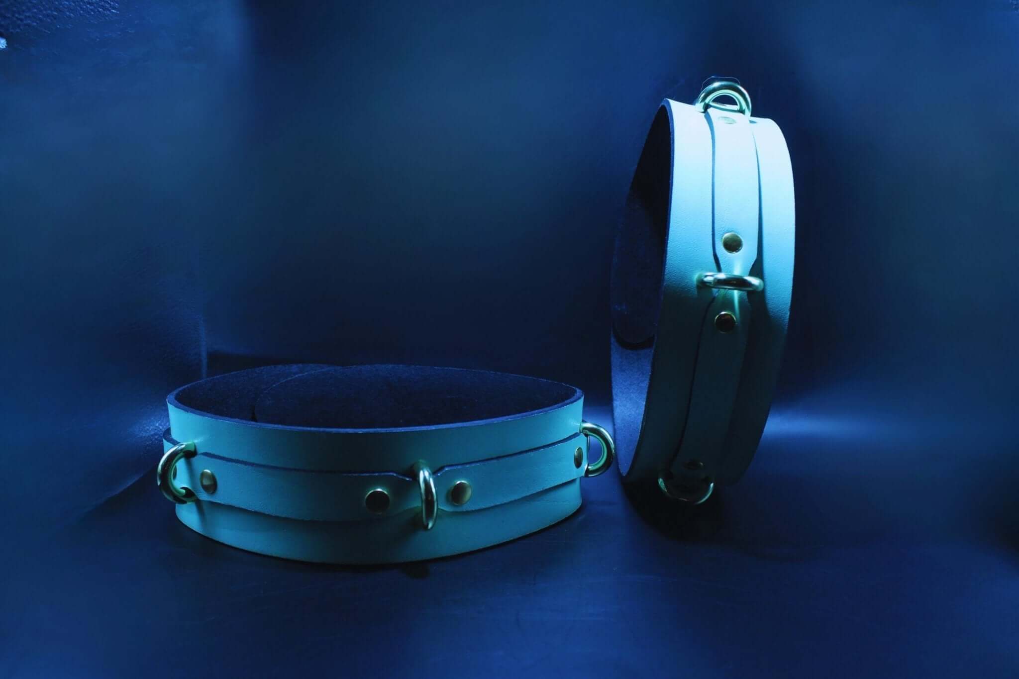 Adjustable leather thigh cuffs in blue light, ideal for bondage gear and BDSM play, featuring metal hardware and stylish design.