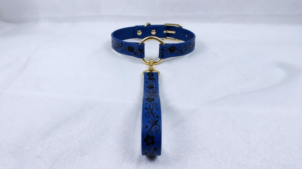 A blue collar with a gold buckle and a black floral pattern on the strap is displayed against a white background.