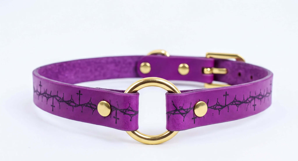 A purple leather collar with a gold-colored buckle and a circular ring design featuring a black cross pattern.