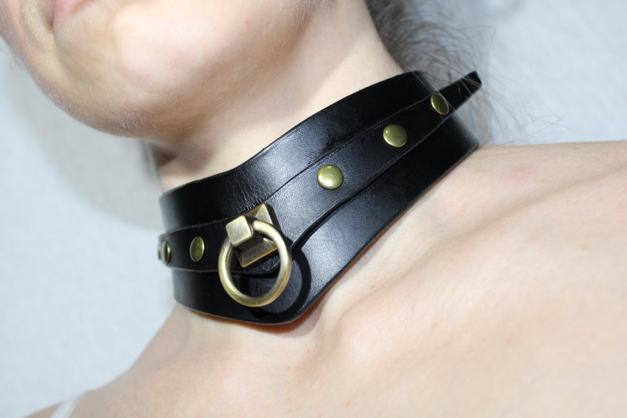 A close-up view of a person's neck wearing a black leather choker necklace with gold-colored studs and a metal ring.