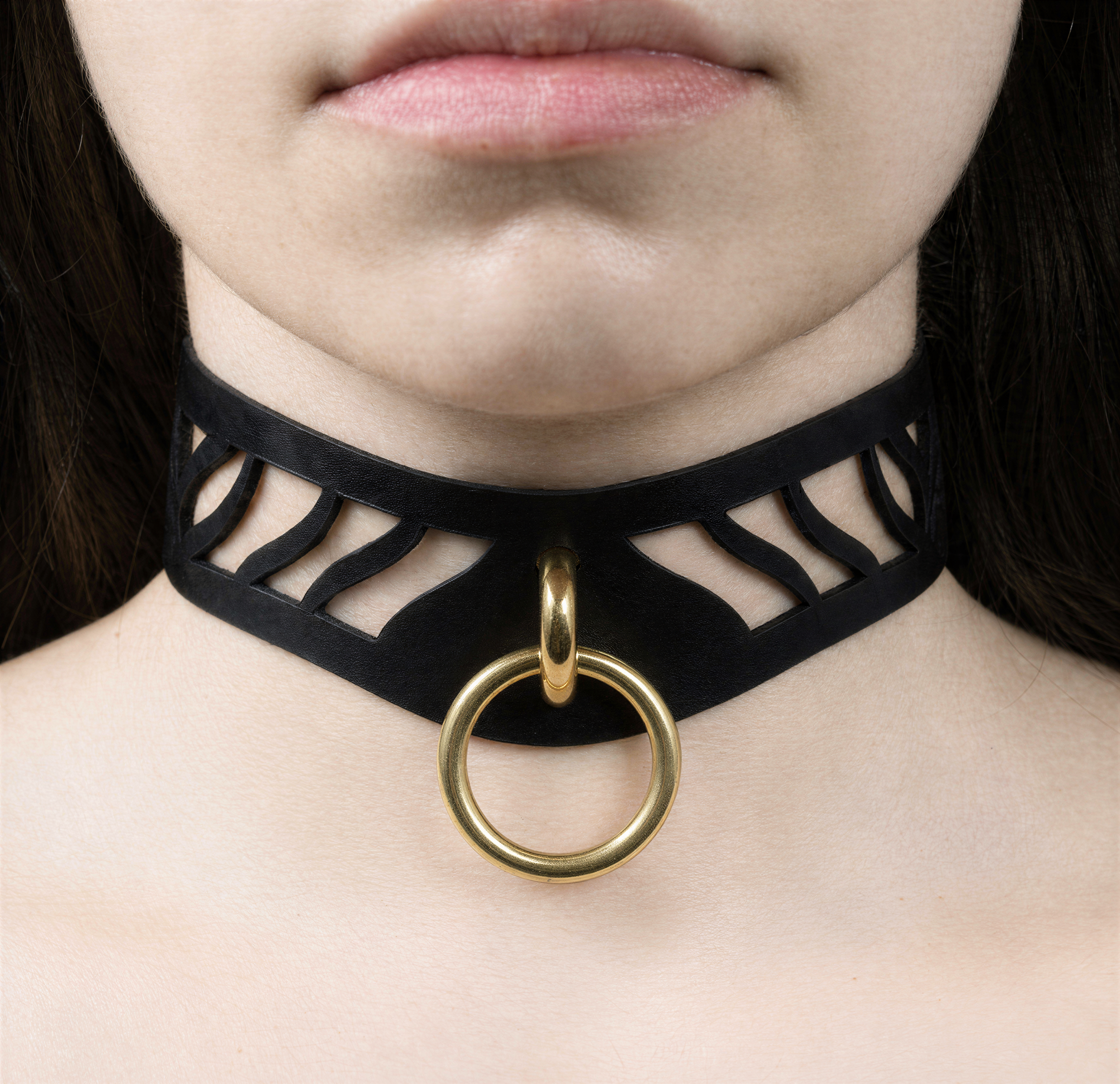A close-up of a person's neck and chest area, featuring a black choker necklace with a gold ring and a cutout design.
