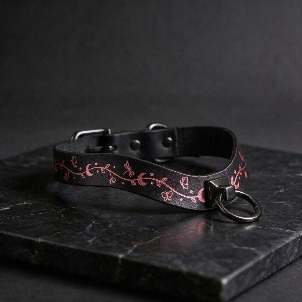 A black leather collar with a pink floral design and a metal ring on the front, resting on a dark surface.