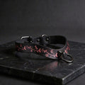 A black leather collar with a pink floral design and a metal ring on the front, resting on a dark surface.